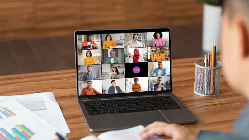 A man sits at a desk in a home office and uses a laptop to participate in a video conference meeting. The laptop screen displays a grid of video windows featuring a diverse group of people