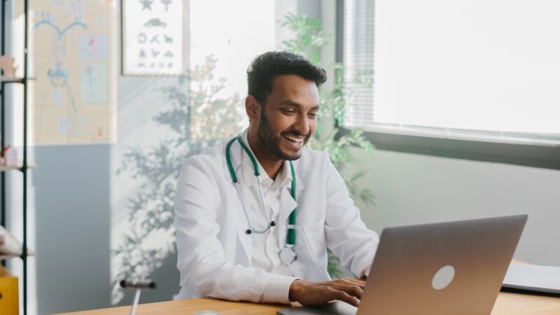 An Asian doctor uses a laptop to conduct an online consultation with a patient.