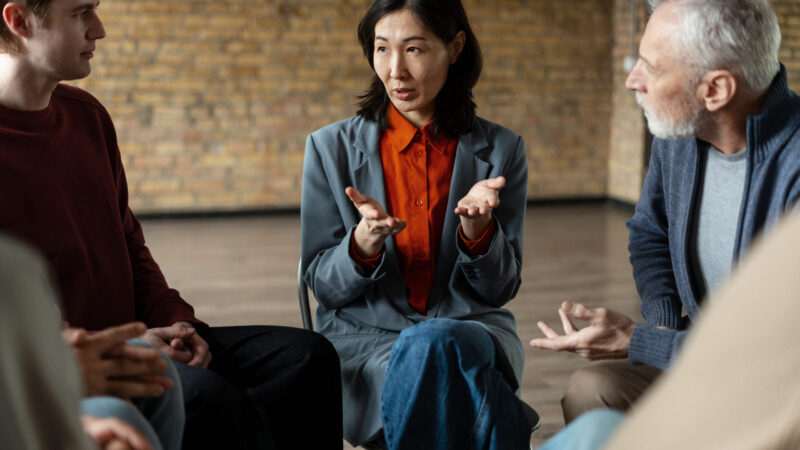 Asian female therapist leading group therapy session, gesturing with hands while speaking to diverse group of patients