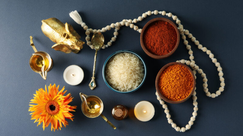 Bowls with spices and rice, copper utensils, candles, beads and an elephant figurine on blue background, top view