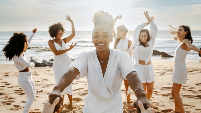 Diverse group of happy young female friends having fun and dancing outdoors on the beach, women celebrating hen party on coastline. Enjoying our beach day together