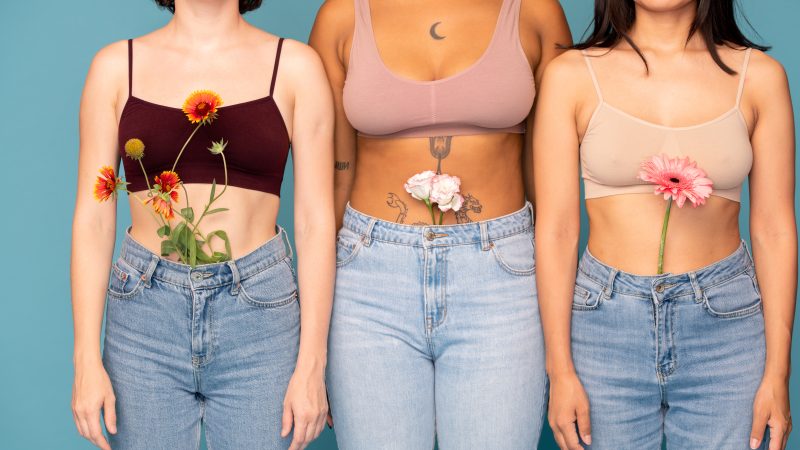 Front view of three young intercultural women with fresh flowers in jeans standing in row close to one another against blue background