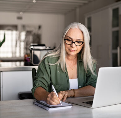 Senior stylish woman taking notes in notebook while using laptop at home. Old freelancer writing details on book while working on laptop in living room. Focused cool lady writing notary in notepad.