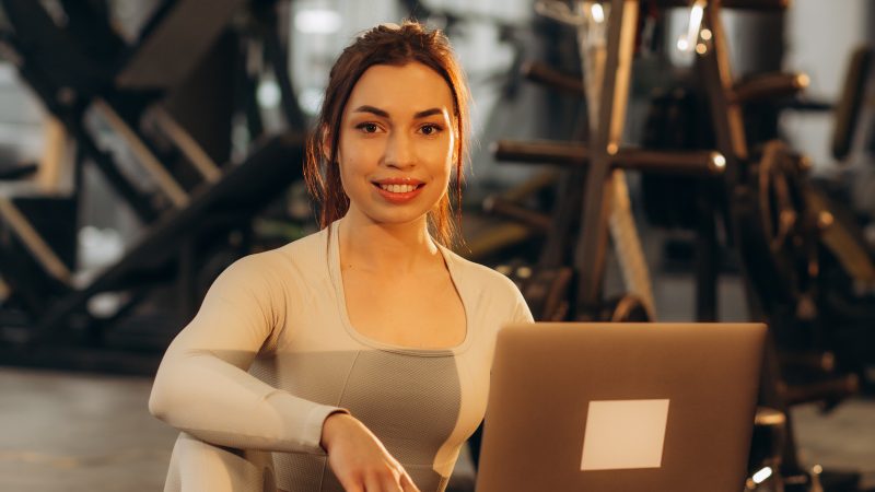 Smiling girl with laptop in a gym. Athlete with dumbbells on a blurred background. Active lifestyle concept.