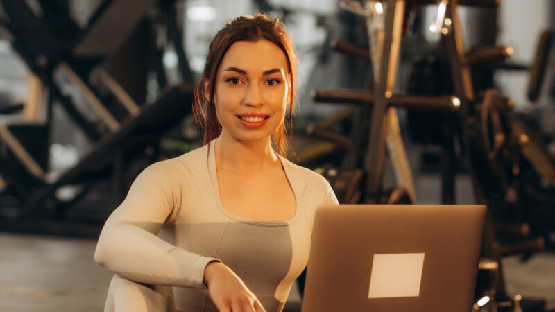 Smiling girl with laptop in a gym. Athlete with dumbbells on a blurred background. Active lifestyle concept.
