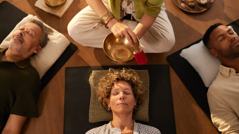 Top view of sound healing session at indoor wellness retreat. Holistic therapist using singing tibetan bell while group lying on back in deep relaxation. High angle view of peaceful moment of energy work, surrounded by crystals and calming atmosphere for people to meditate.