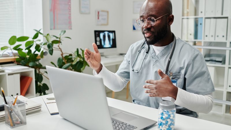 Young confident African American online doctor in uniform sitting by desk in front of laptop in clinics and giving medical advice to patient