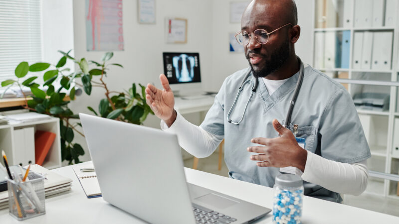 Young confident African American online doctor in uniform sitting by desk in front of laptop in clinics and giving medical advice to patient